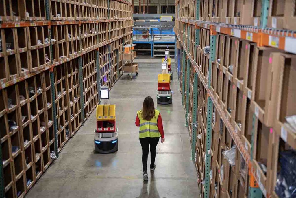 Woman Walking Down Warehouse Aisle With Robots